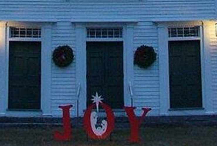 Three doors in the exterior of a white building. Wreaths hang between the doors. In front of the building, red letters spelling "Joy" are staked into the ground. A nativity scene is depicted inside the O.