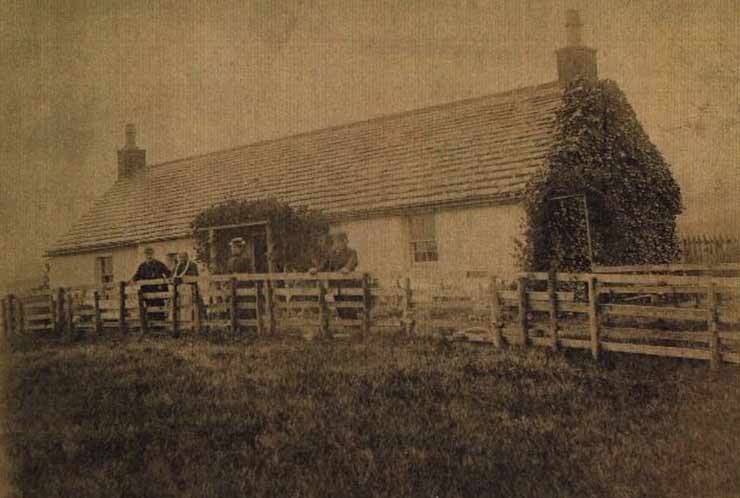 A historical photo of 4 people standing in front of a house and behind a wooden fence